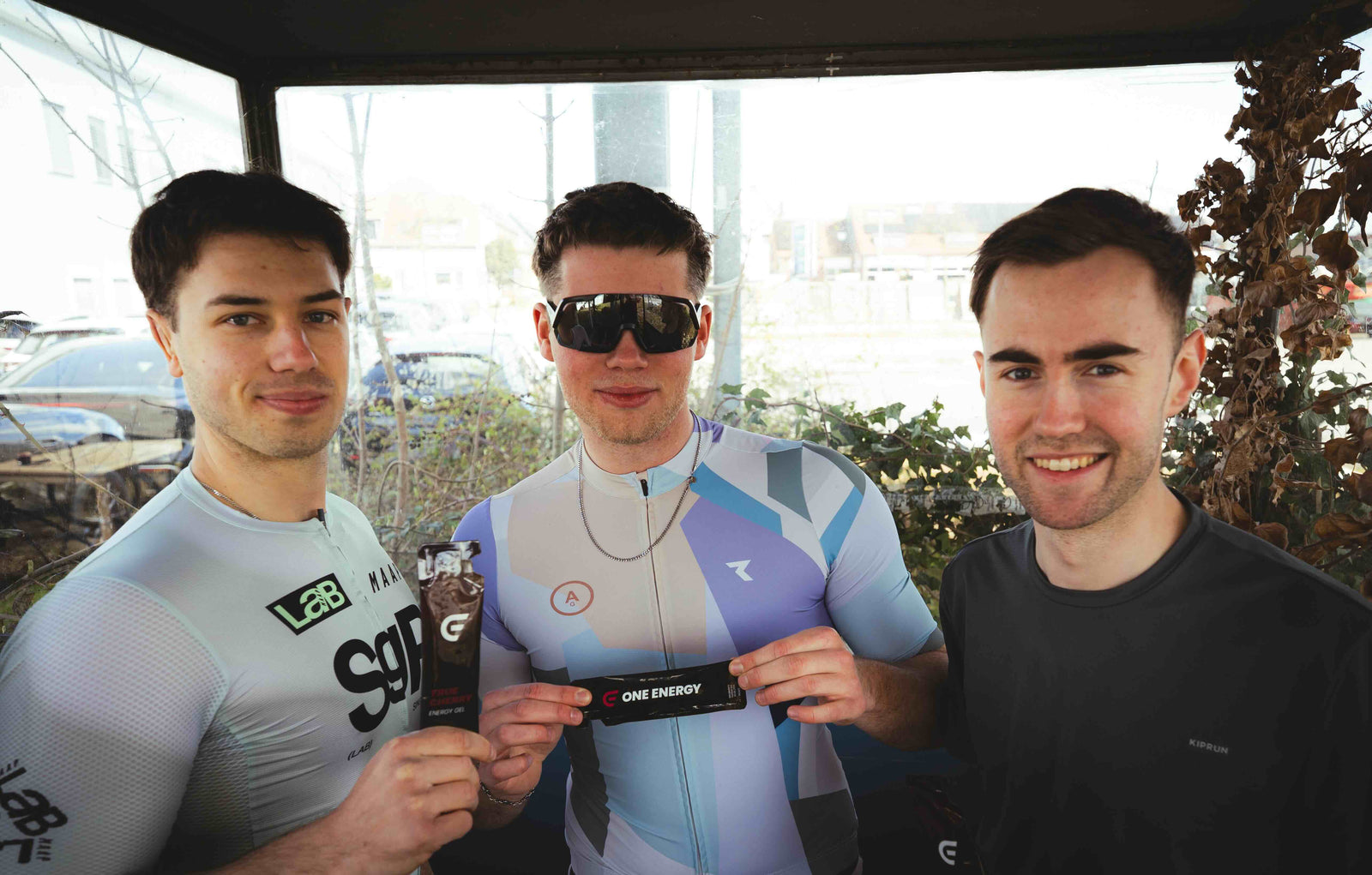Die drei Gründer von ONE ENERGY lächeln in die Kamera und präsentieren ihre Energy Gel in Sportbekleidung