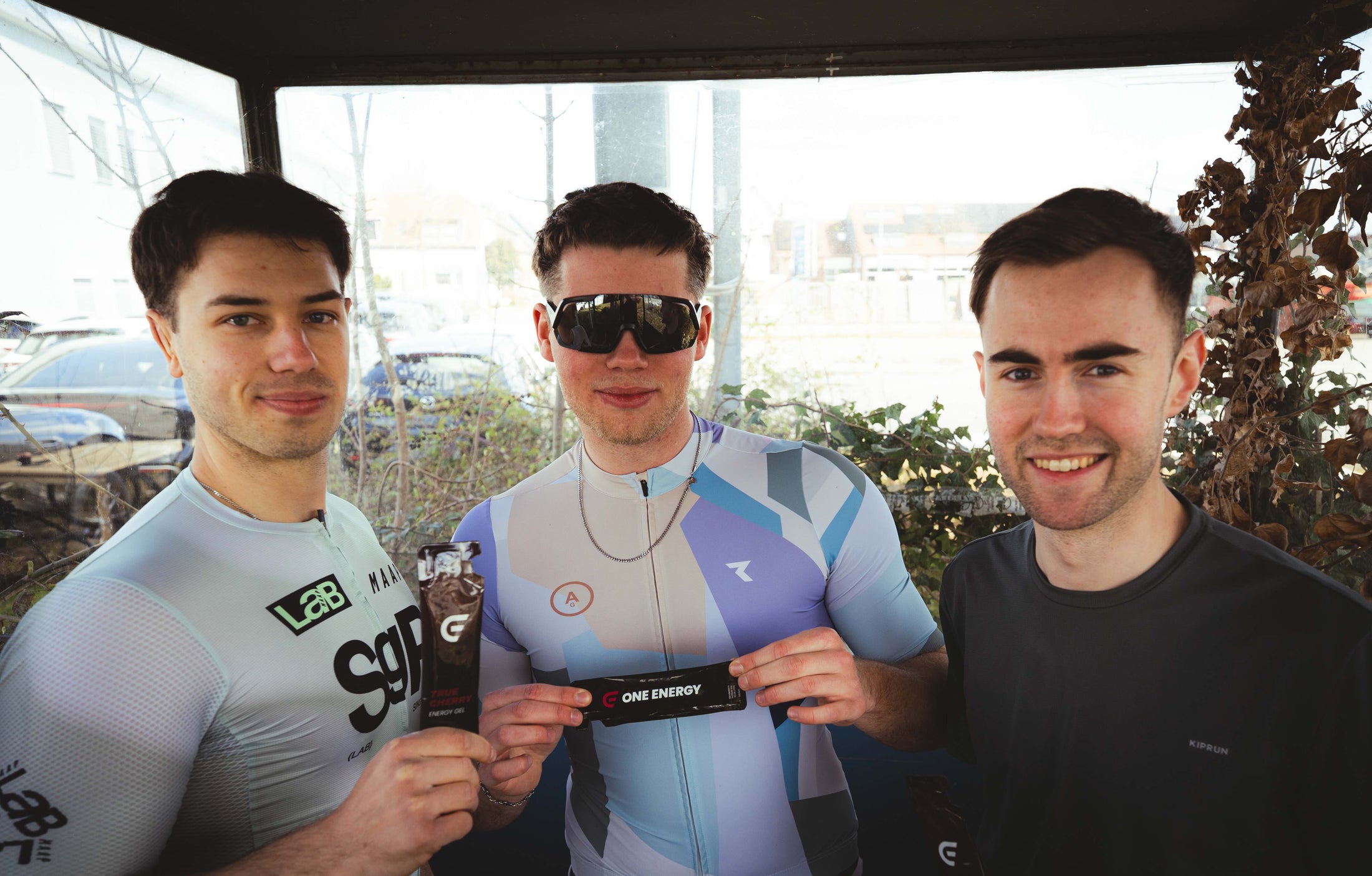 Die drei Gründer von ONE ENERGY lächeln in die Kamera und präsentieren ihre Energy Gel in Sportbekleidung